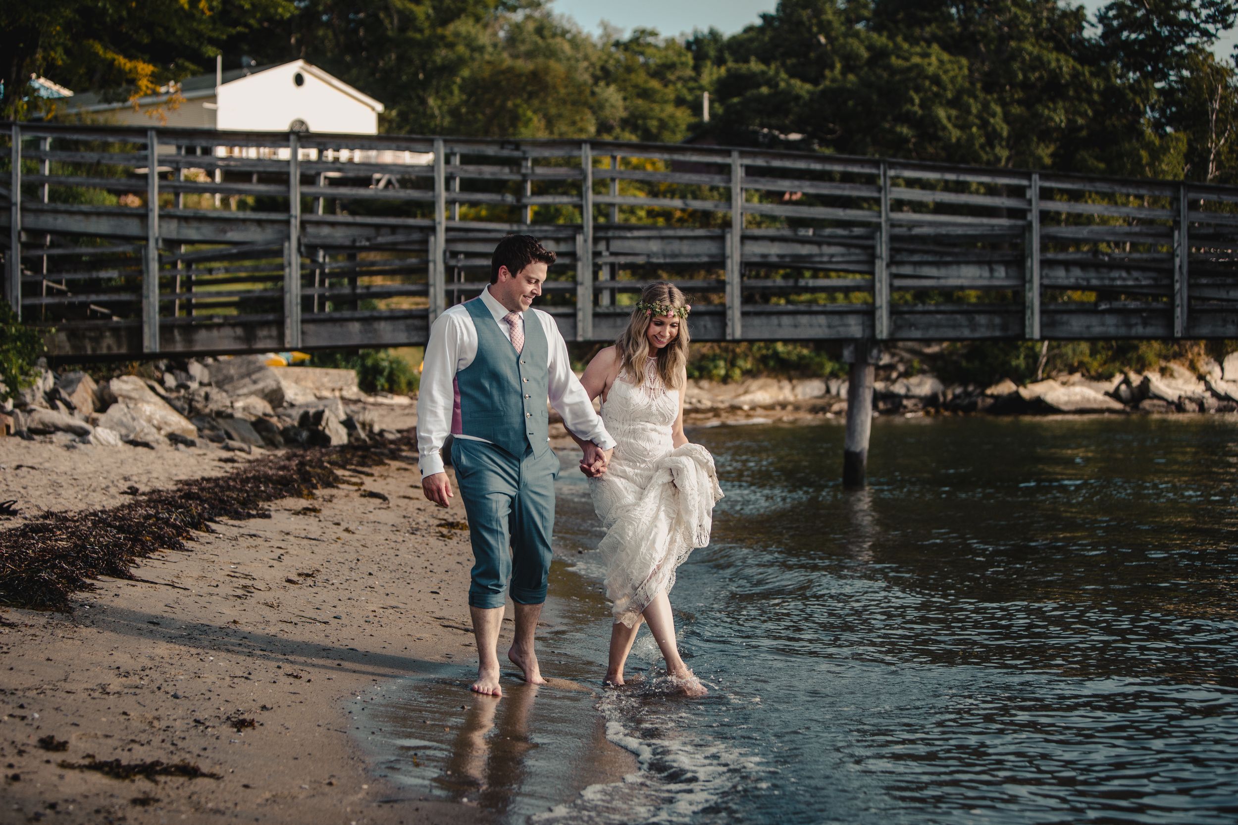 Bride and groom walking barefoot along a sandy shoreline near a wooden dock on their wedding day.