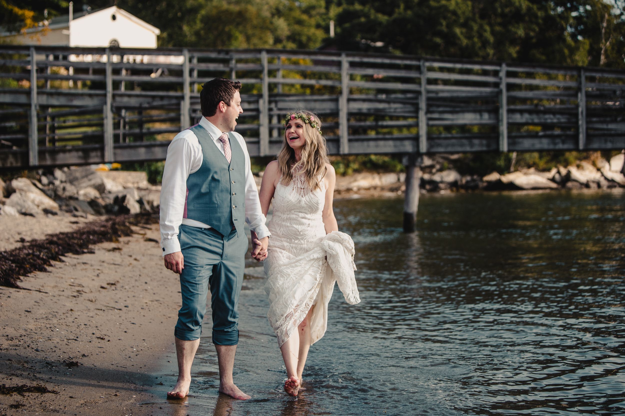 Bride and groom walk hand-in-hand barefoot along a rocky shoreline near a wooden bridge on their wedding day.