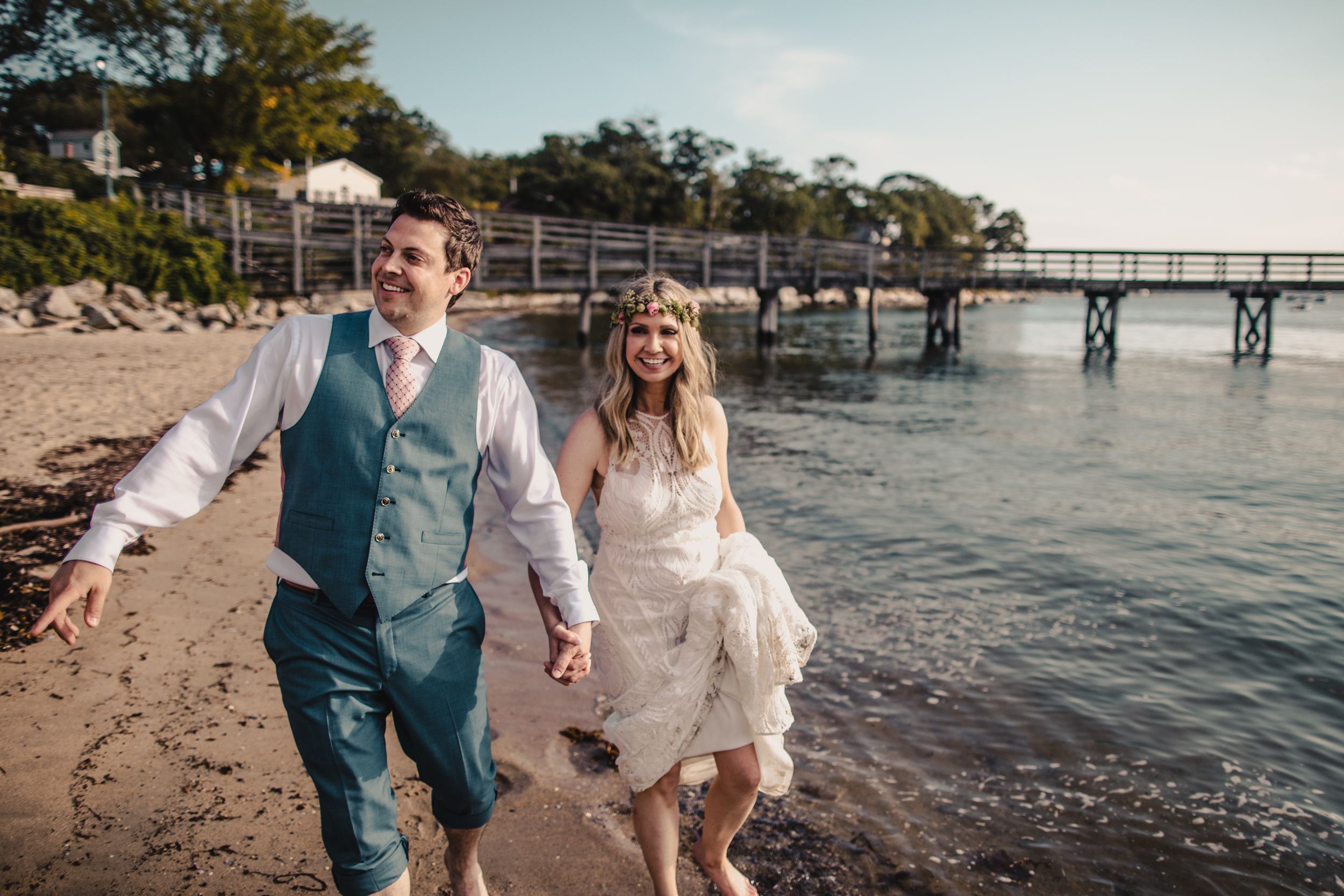 Happy newlyweds walk barefoot on a sandy beach holding hands with a pier visible in the background.