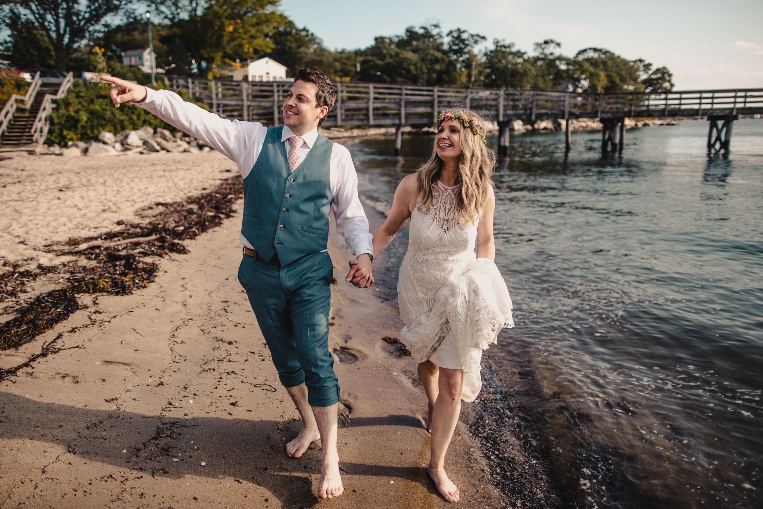 Barefoot bride and groom walk hand-in-hand along sandy beach shoreline near pier on their wedding day.