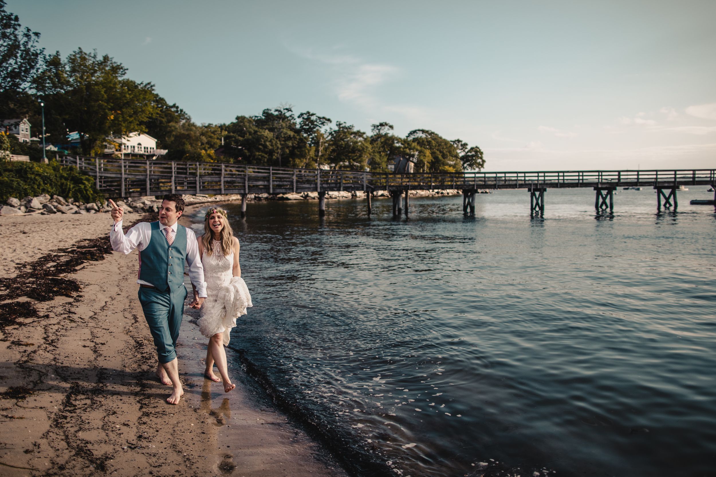Bride and groom walking barefoot along a sandy beach shoreline near a wooden pier at sunset.