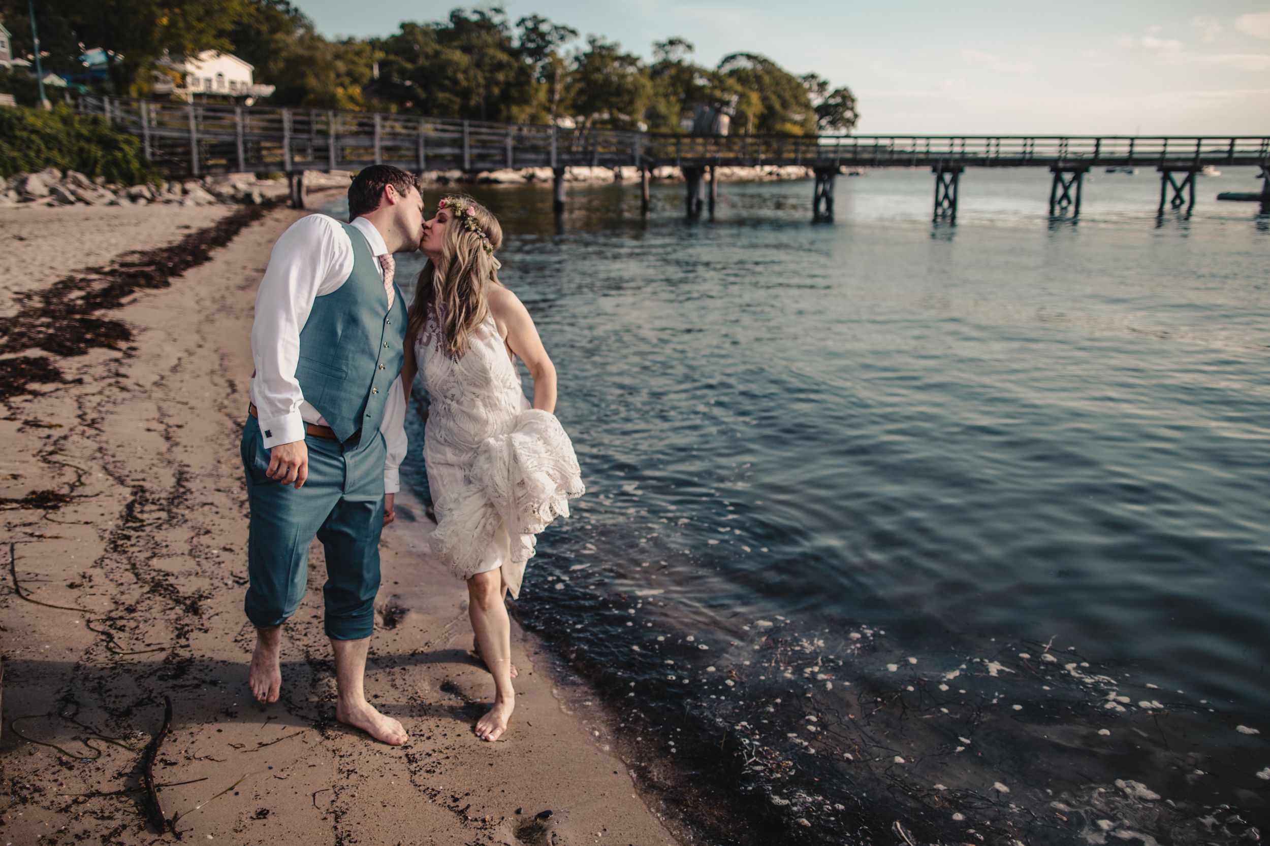 Newlywed couple sharing a romantic kiss on a sandy beach with a wooden pier in the background.