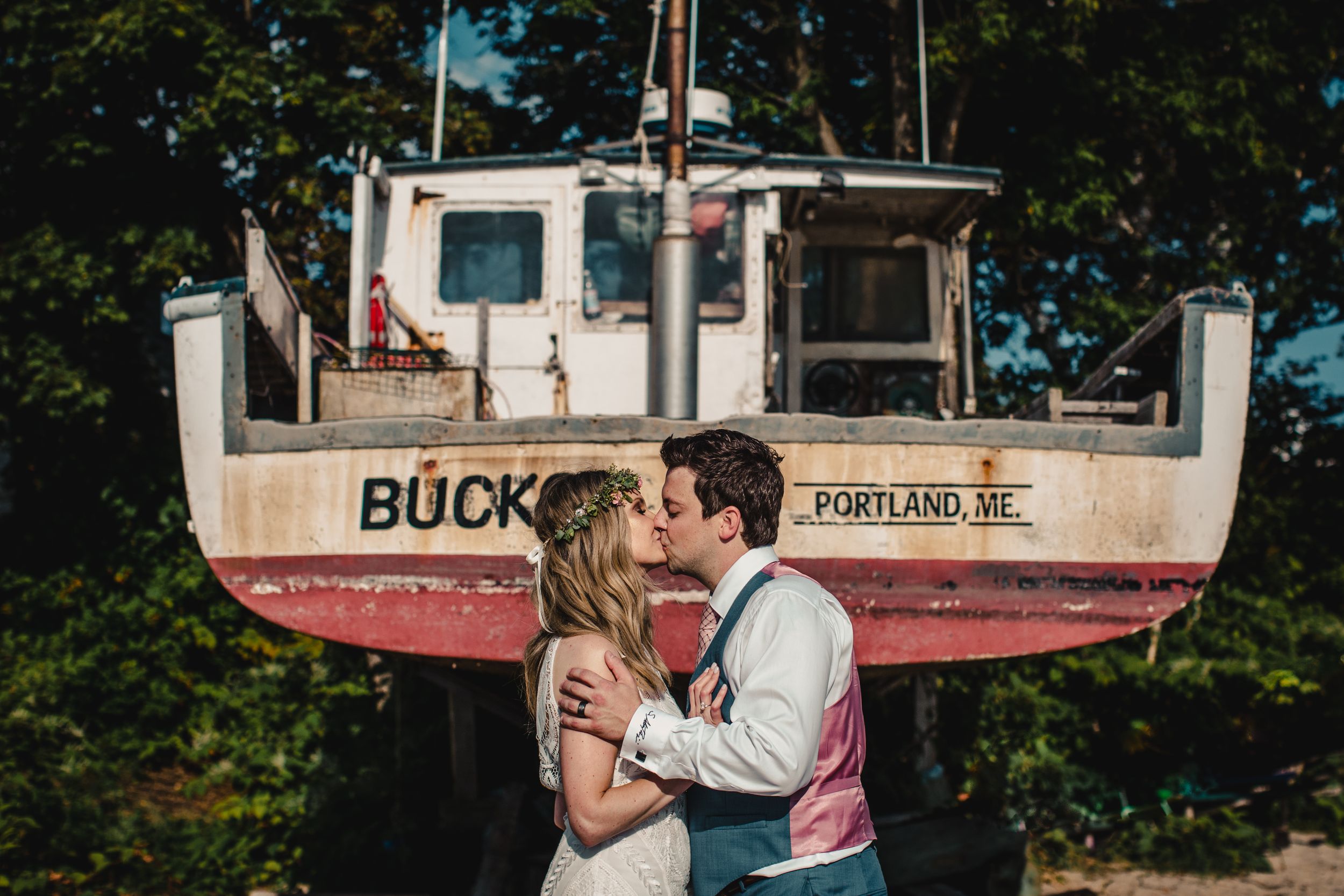 Bride and groom share a romantic kiss in front of a vintage red boat named Buck in Portland, ME.