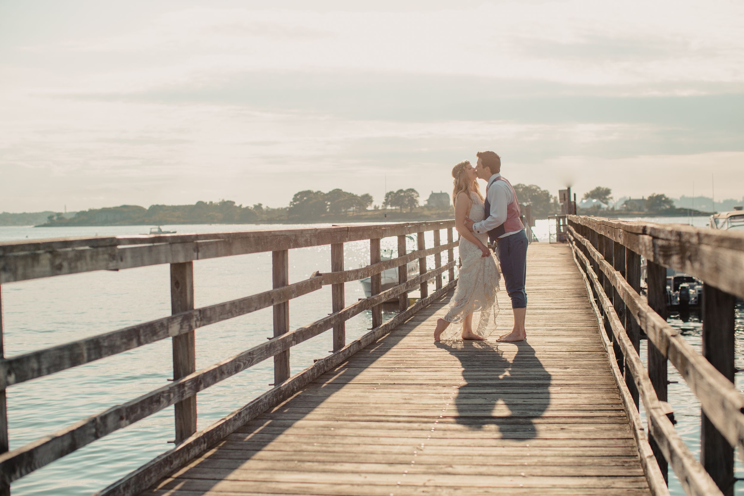 Couple sharing a romantic kiss on a wooden pier overlooking calm blue water on a sunny day.