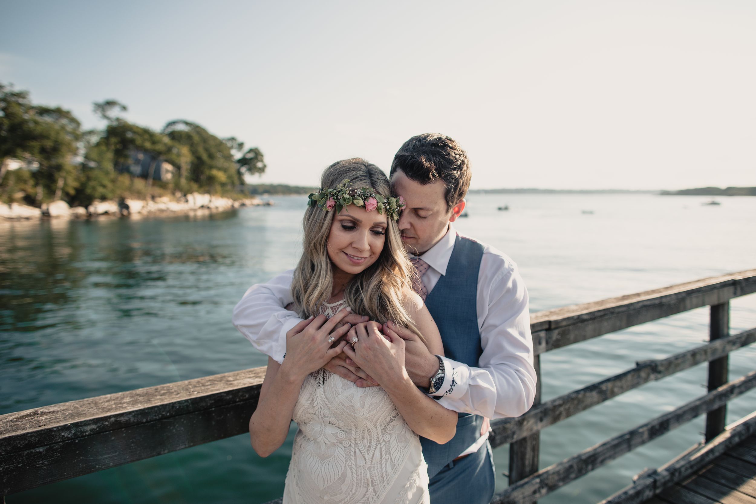 Groom embraces bride from behind on wooden pier overlooking turquoise water on their wedding day.