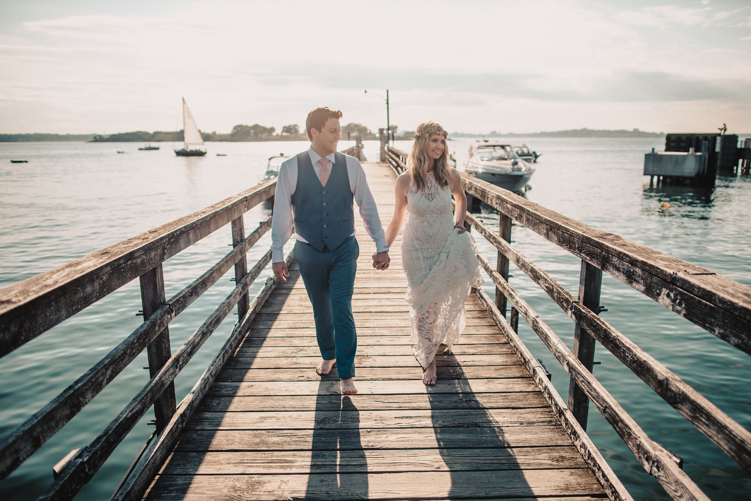 Bride and groom holding hands walking on a wooden pier at sunset with sailboats in the background.