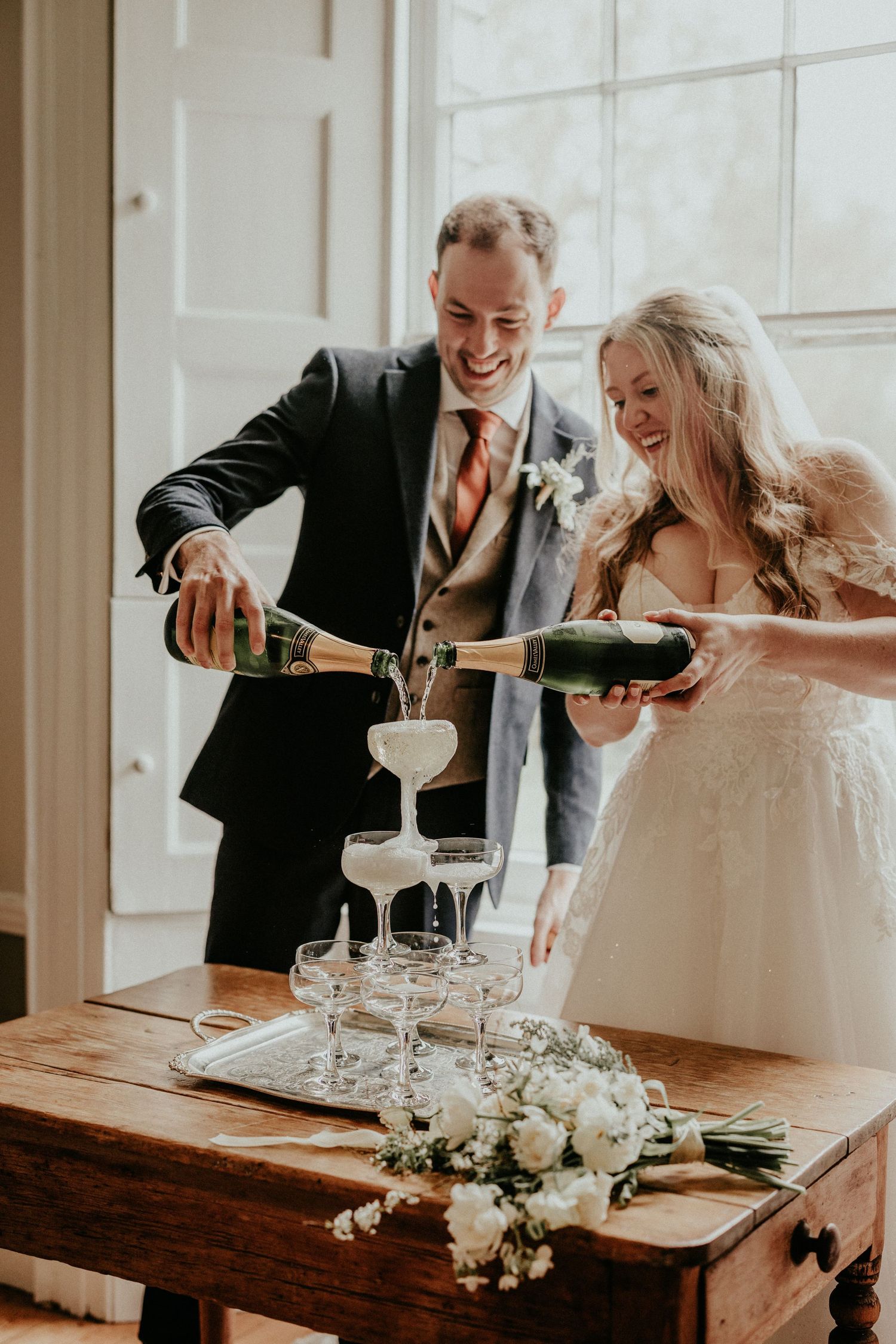 Smiling bride and groom pouring champagne tower at wooden table with white flowers at their wedding reception.