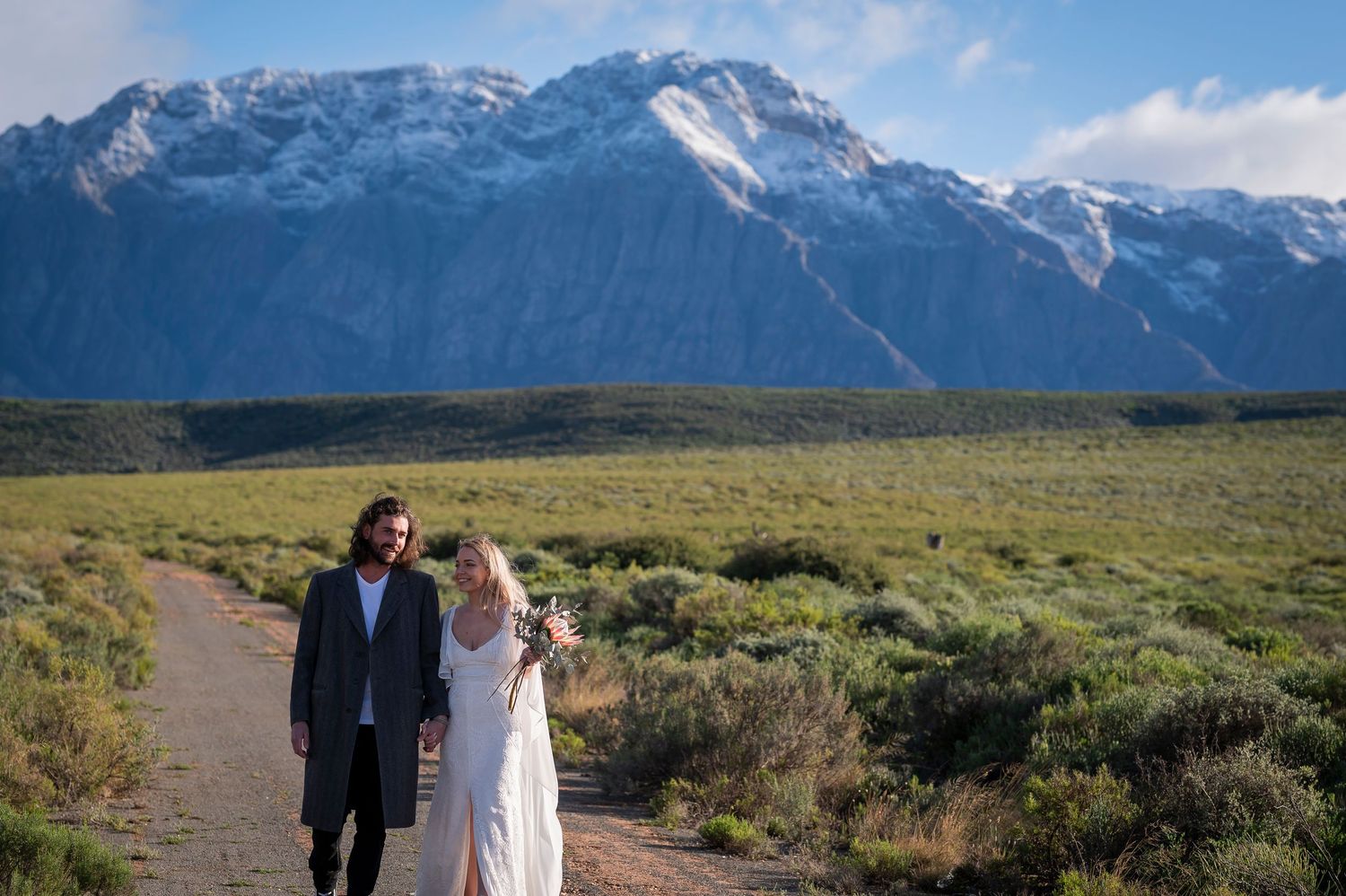 Couple walking hand in hand on a dirt path with snow-capped mountains and green meadows in the background.