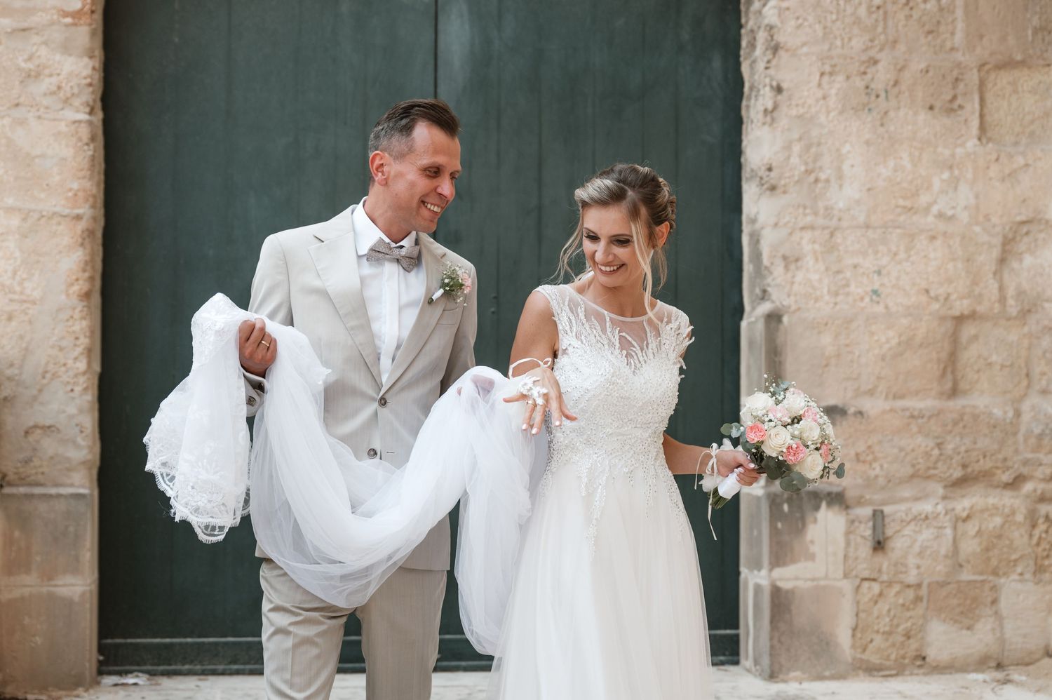 A wedding party stands in front of a rustic stone wall and green door in elegant white attire.
