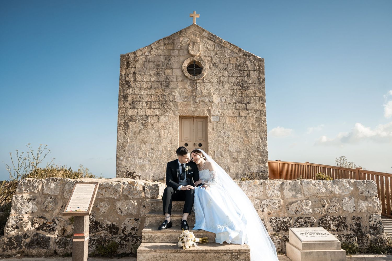 A stone church exterior with a cross on top stands against a bright blue sky in a panoramic view.