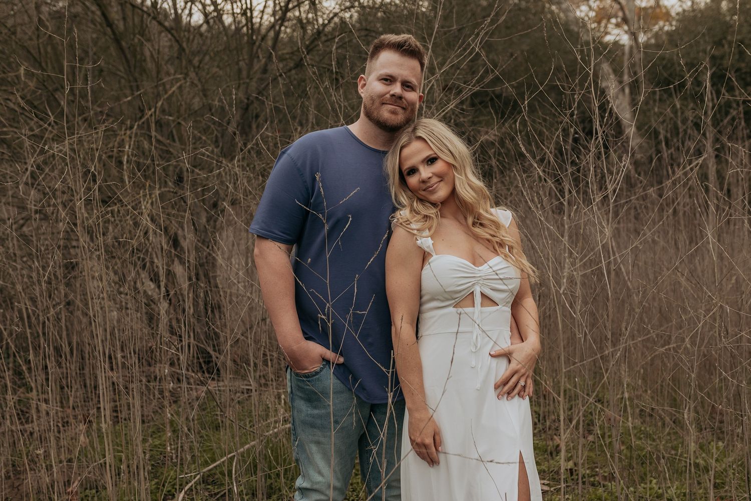 A couple poses together in a rustic outdoor setting surrounded by bare winter trees and brush in San Diego.