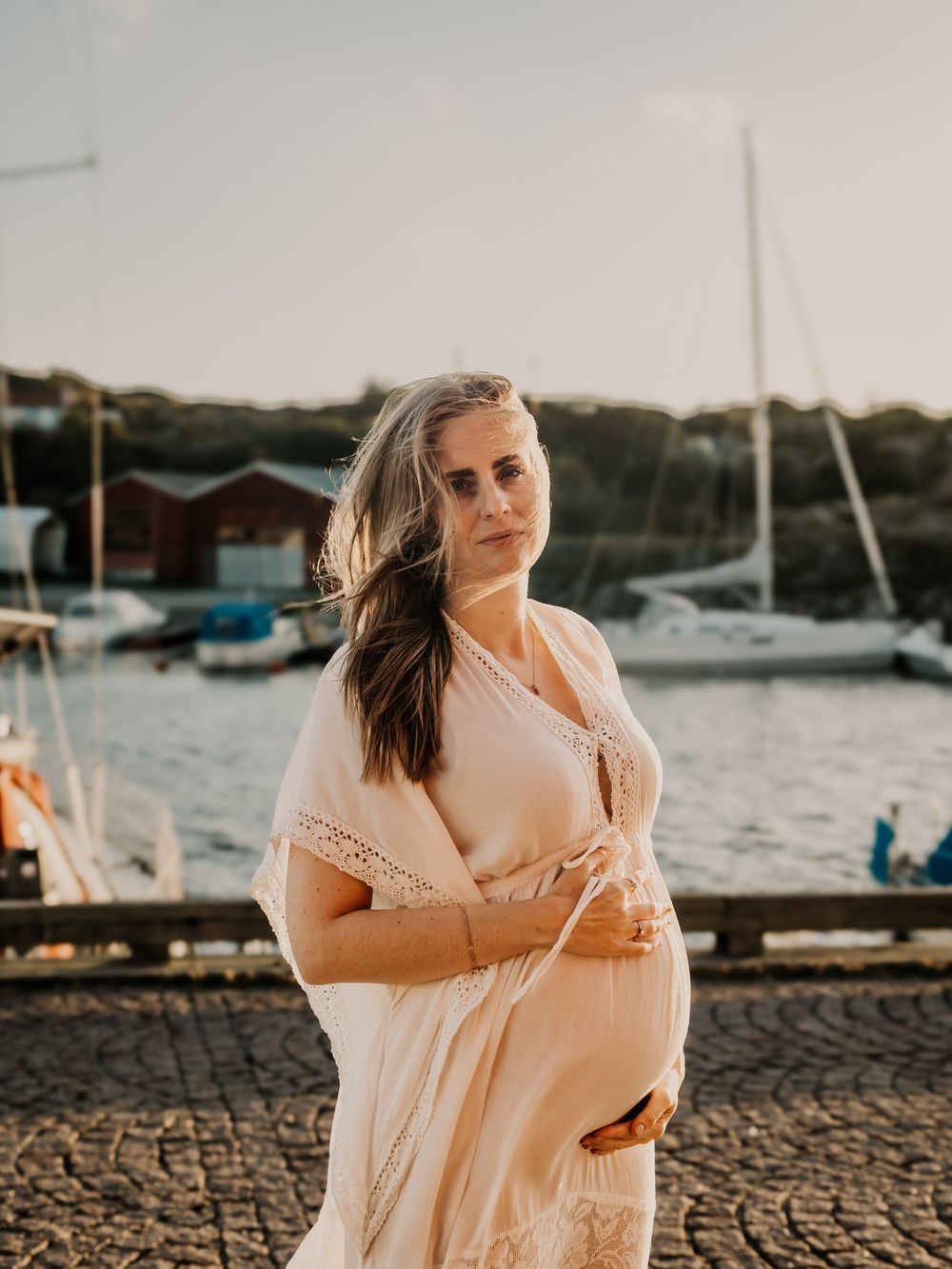Woman in flowing white dress poses gracefully by marina at sunset with boats and water in background.