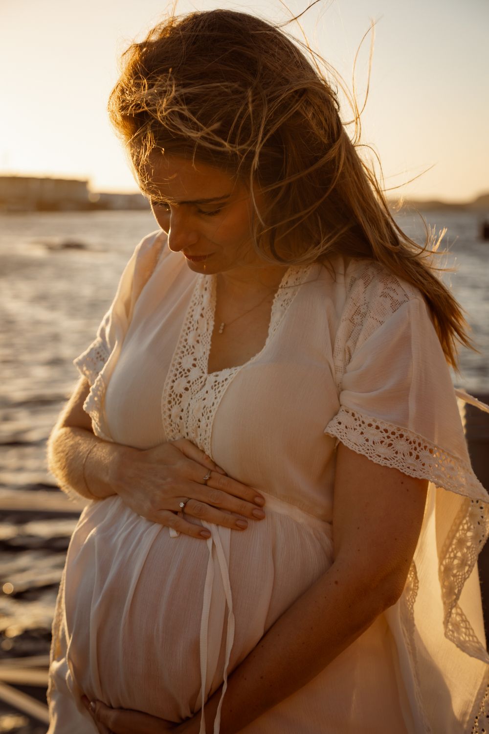 Pregnant woman in white lace dress cradles her belly during sunset beach photoshoot.