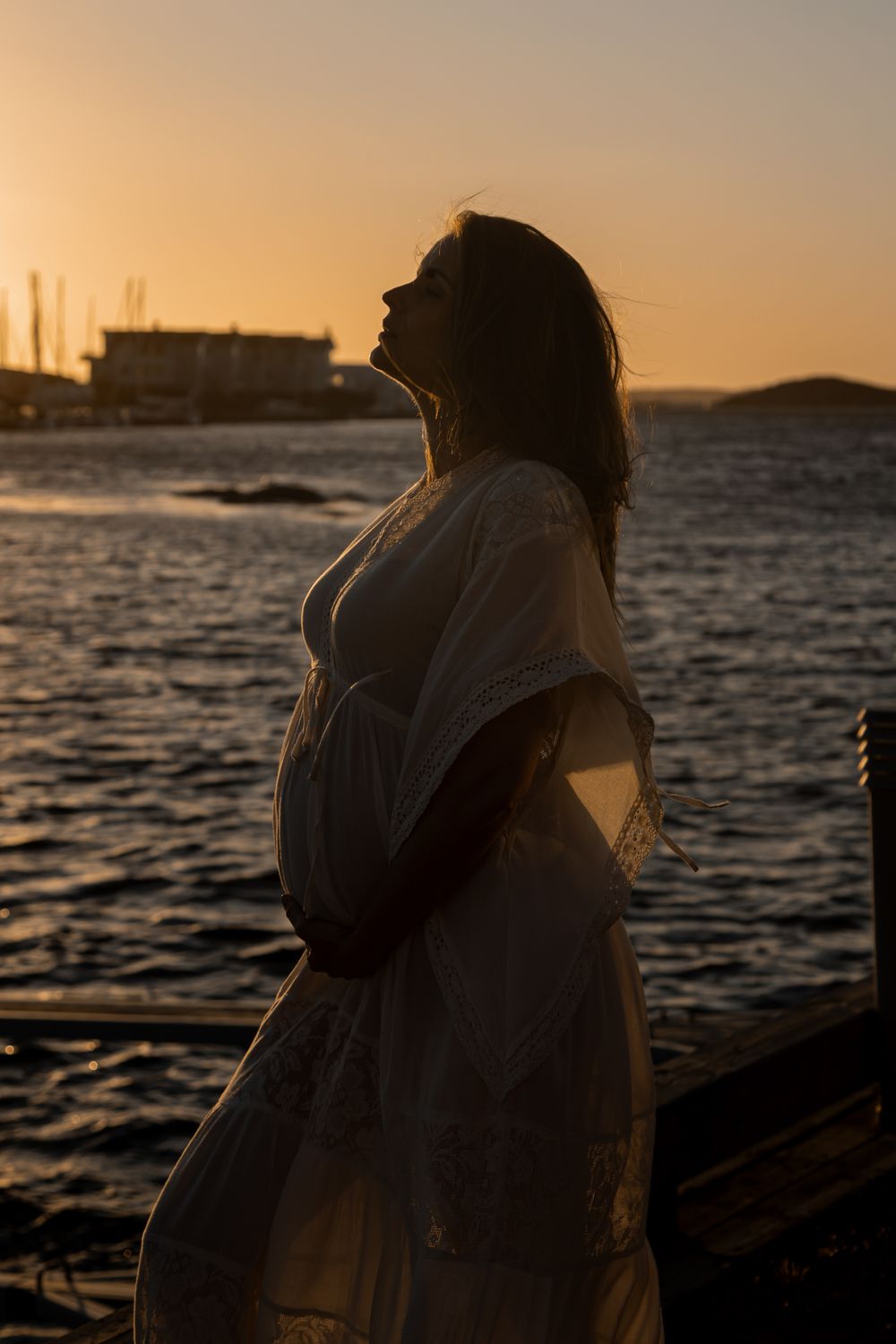 A silhouette stands by the water during golden hour sunset, wearing a flowing white dress against a maritime backdrop.