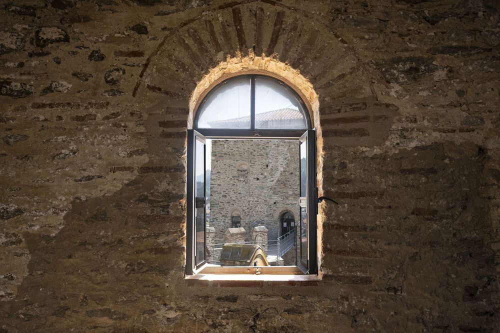Arched stone window in ancient wall reveals silhouetted figures through glass pane framed by rustic masonry.