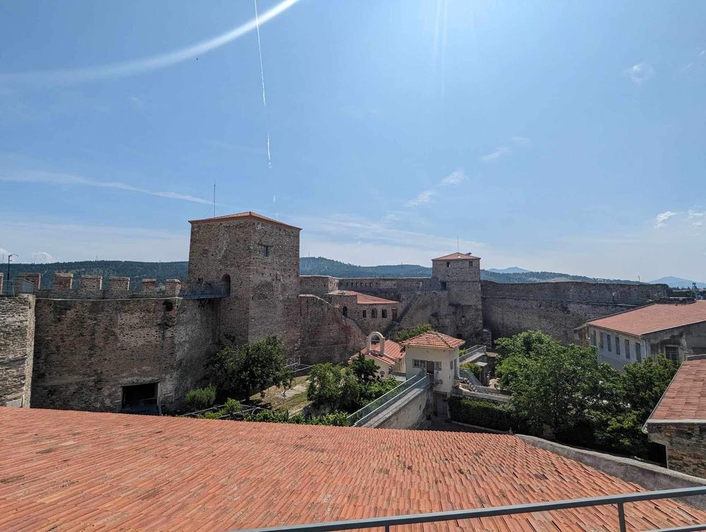 Ancient stone fortress walls and red-tiled roofs stretch across a hilltop under bright blue Mediterranean skies.