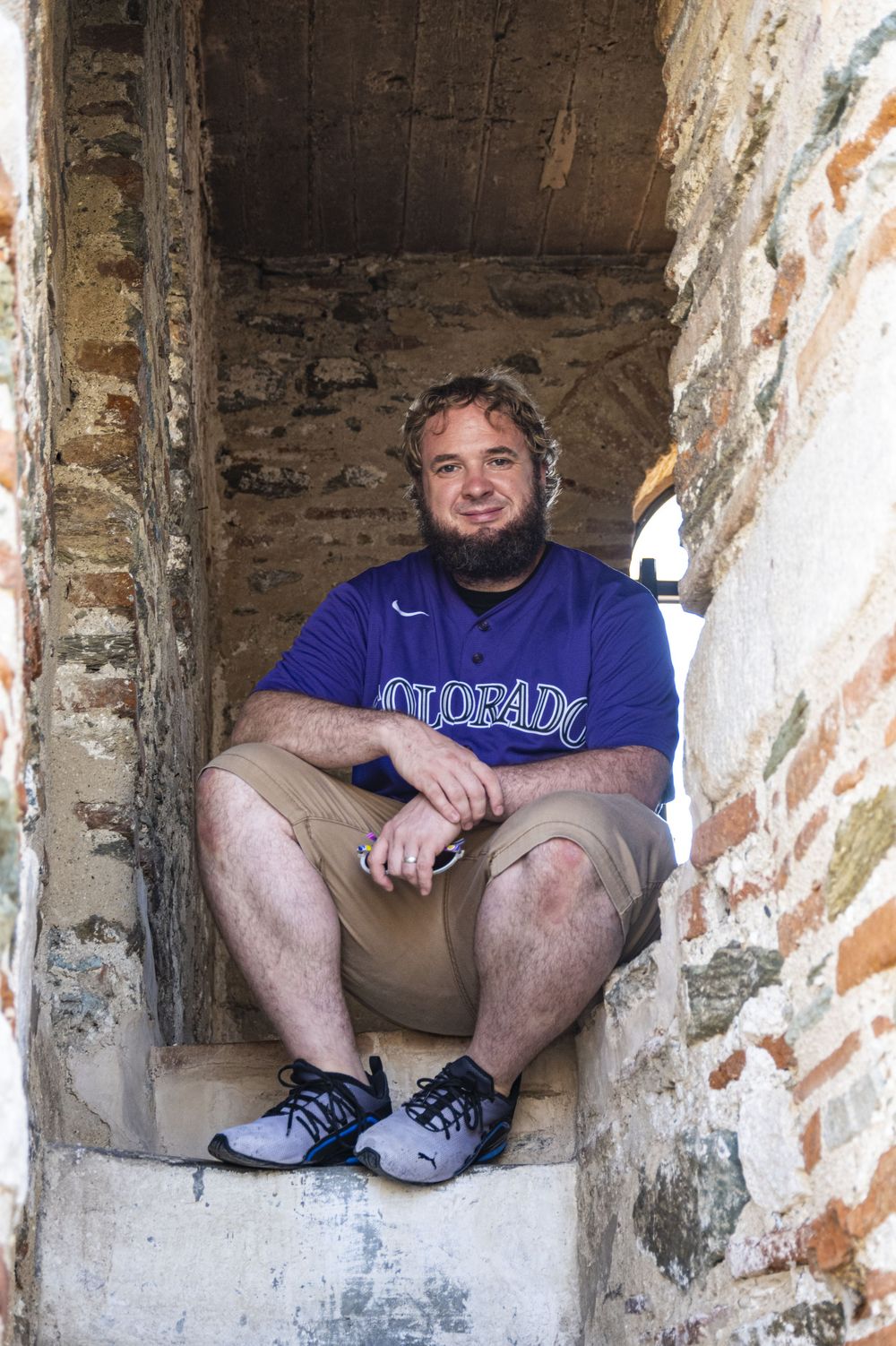 A person sits in an old stone window opening wearing a purple Colorado sweatshirt and khaki shorts.