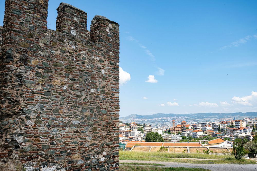 Ancient stone fortress walls stand against a bright blue sky with distant mountains and buildings visible in the background.