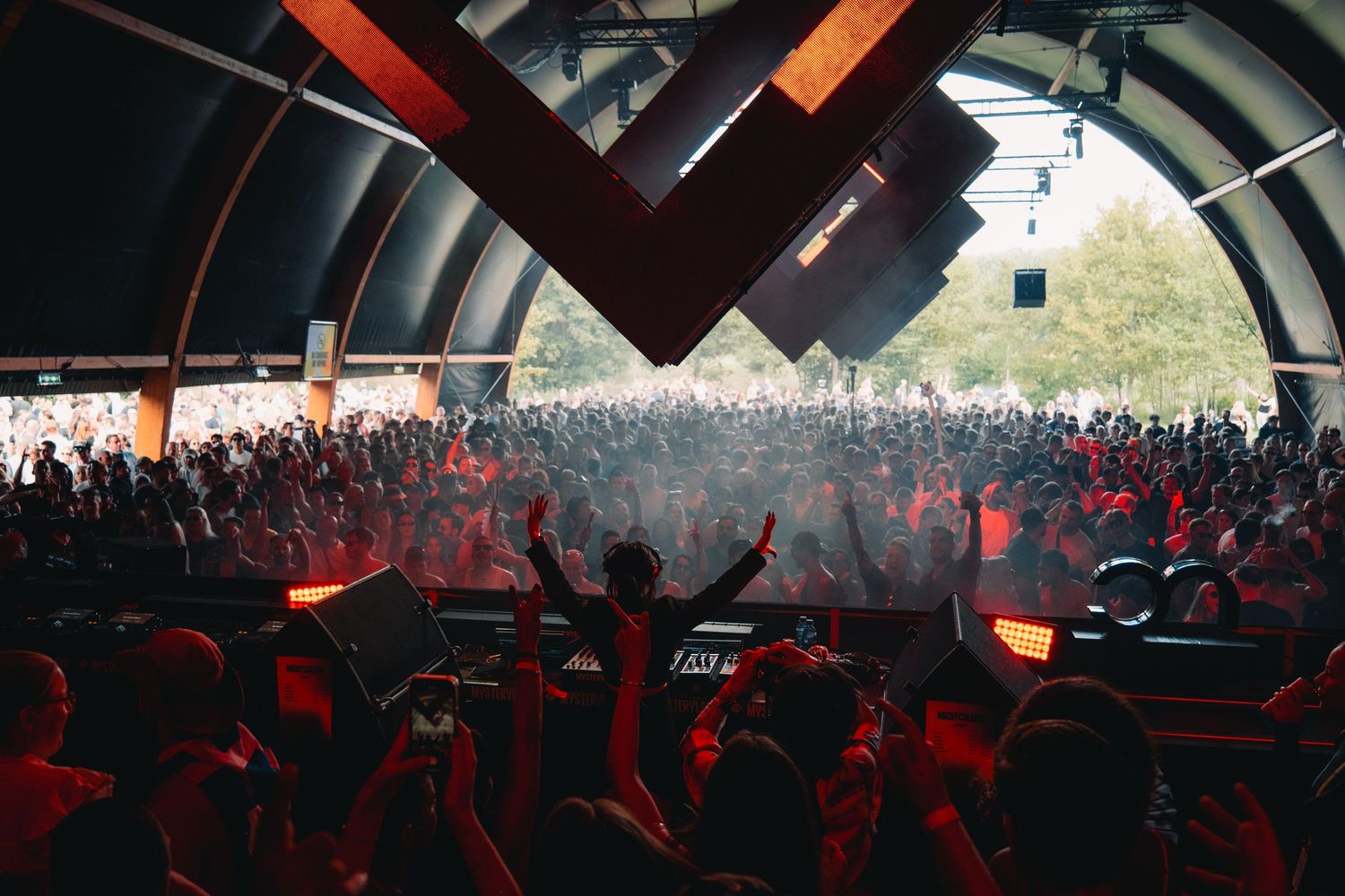 DJ performing on stage bathed in red light before a massive crowd at an outdoor music festival.