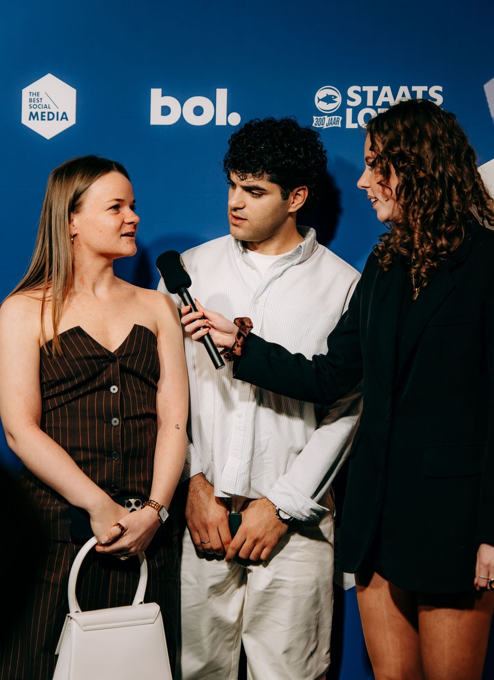 Two women and a man being interviewed on a blue branded press carpet at an event.