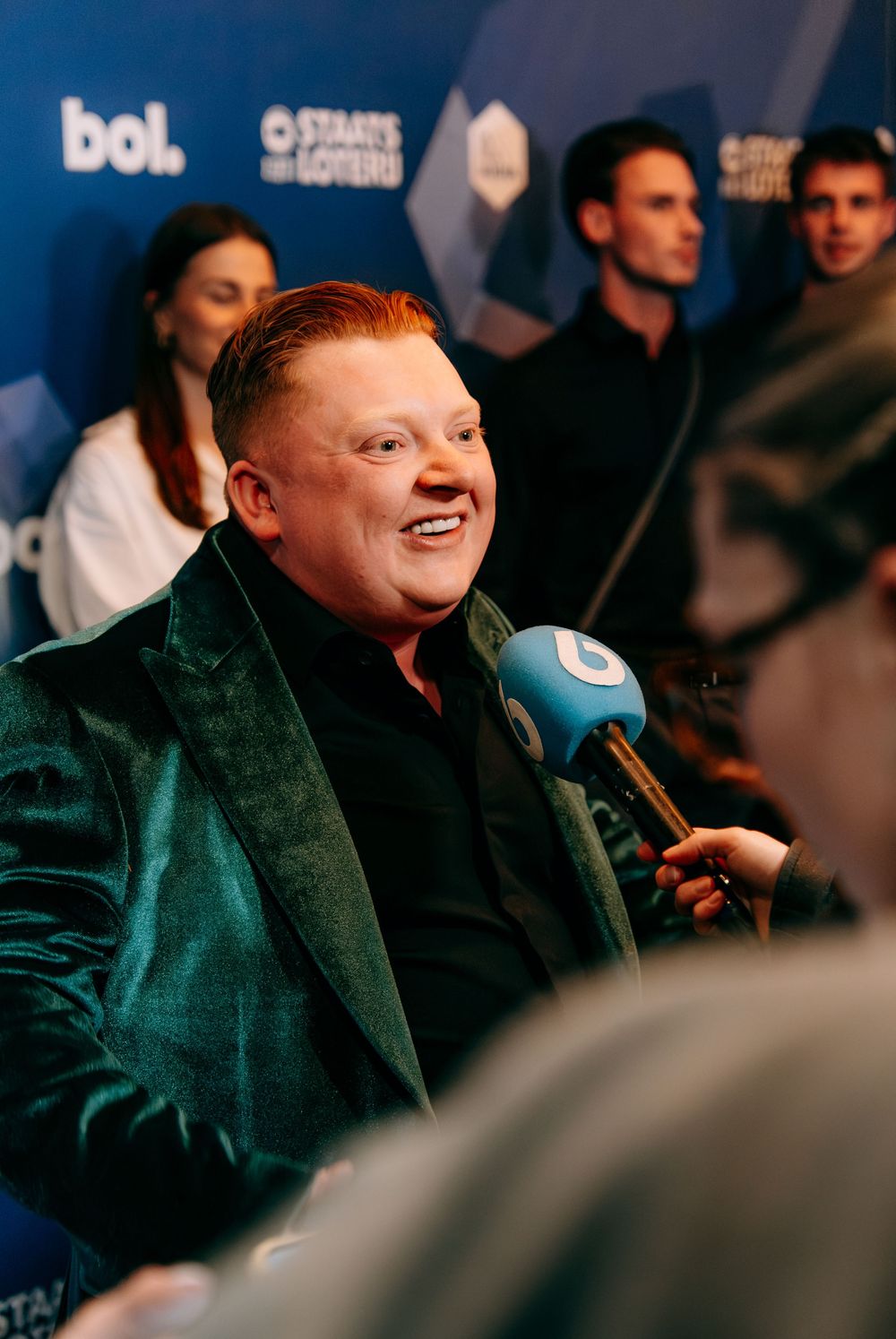 Smiling man in a green velvet blazer being interviewed at a branded press event.