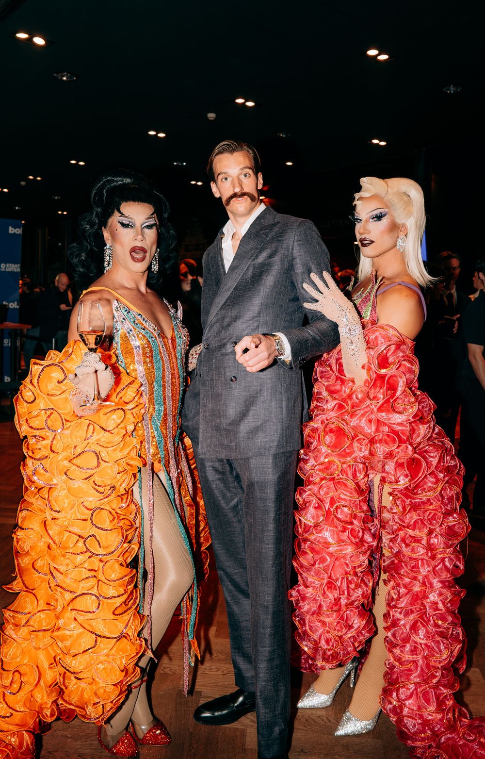 Two drag queens in colorful ruffled gowns posing with a suited man at a gala.