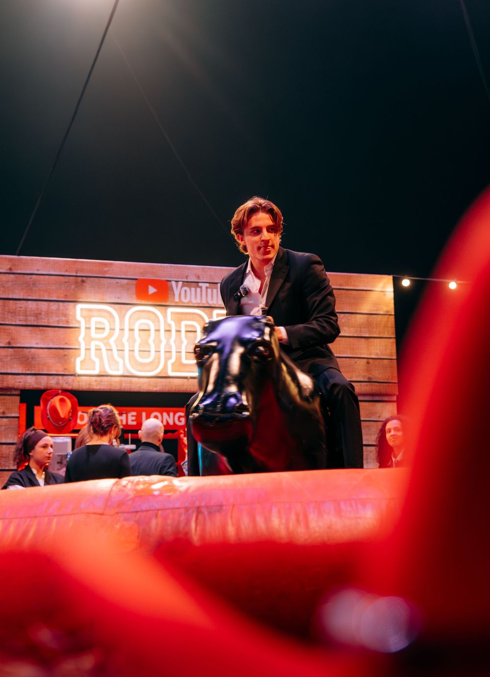 Young man in suit riding mechanical bull at YouTube Rodeo event with wooden backdrop and neon signage.