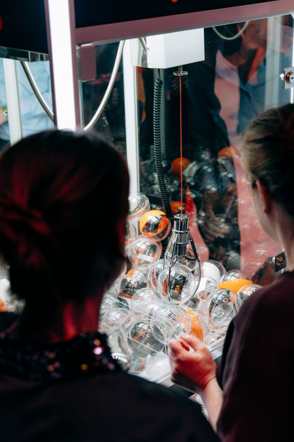 Two people playing claw machine arcade game filled with clear plastic ball capsules at event.