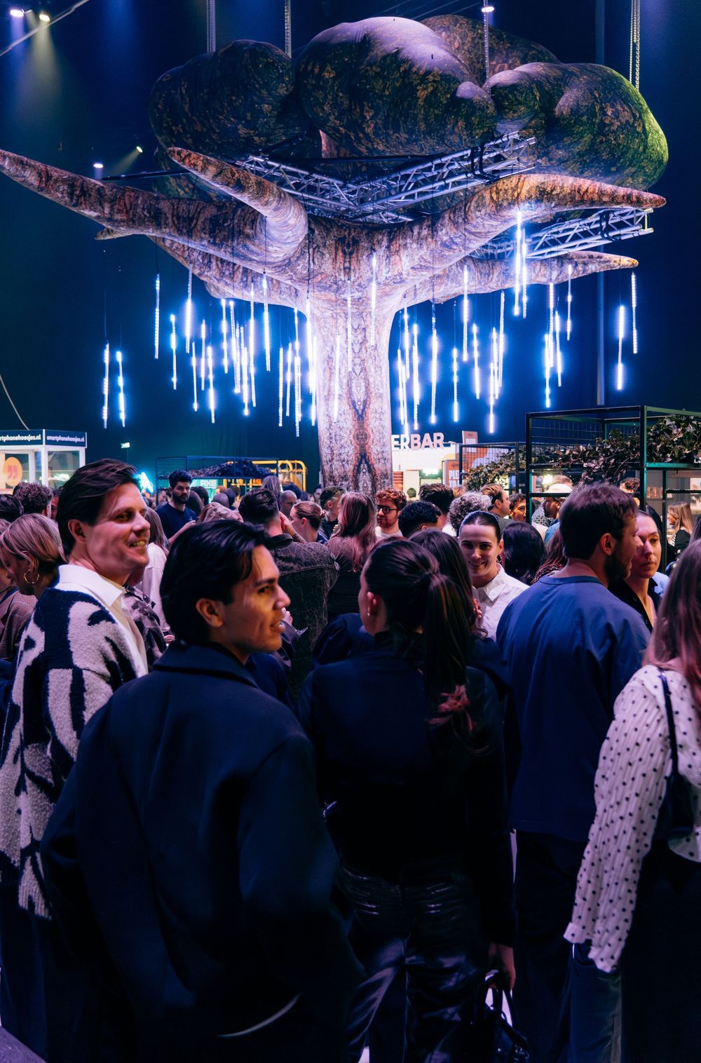 Crowd of people gathered under large illuminated decorative tree installation at indoor entertainment event.