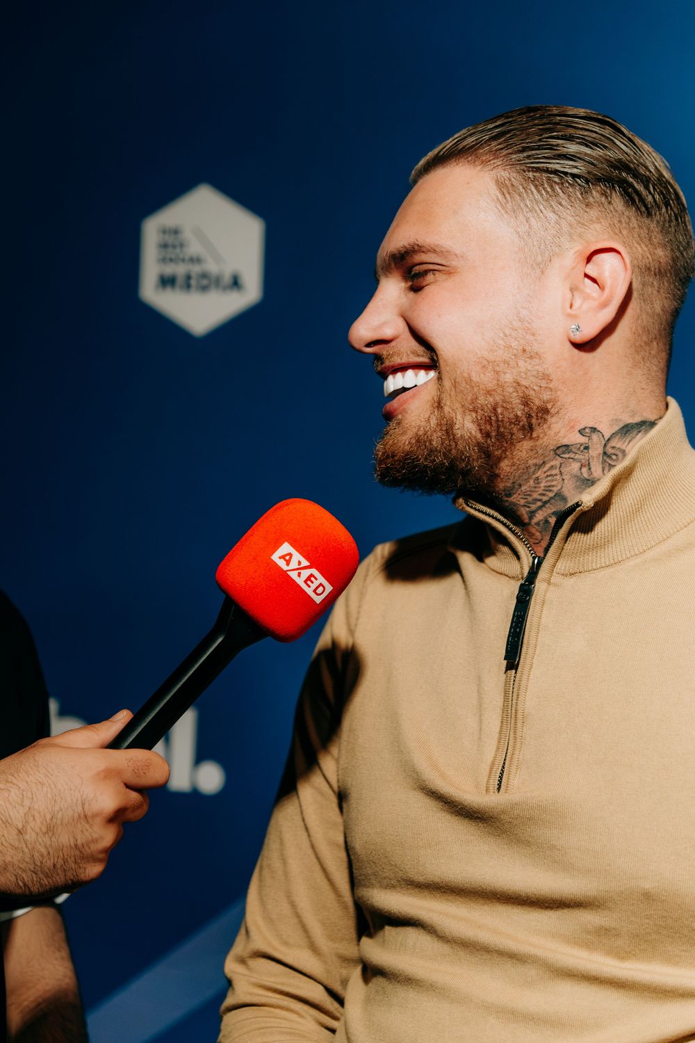 Tattooed man in beige sweater smiling during red microphone interview at media event.