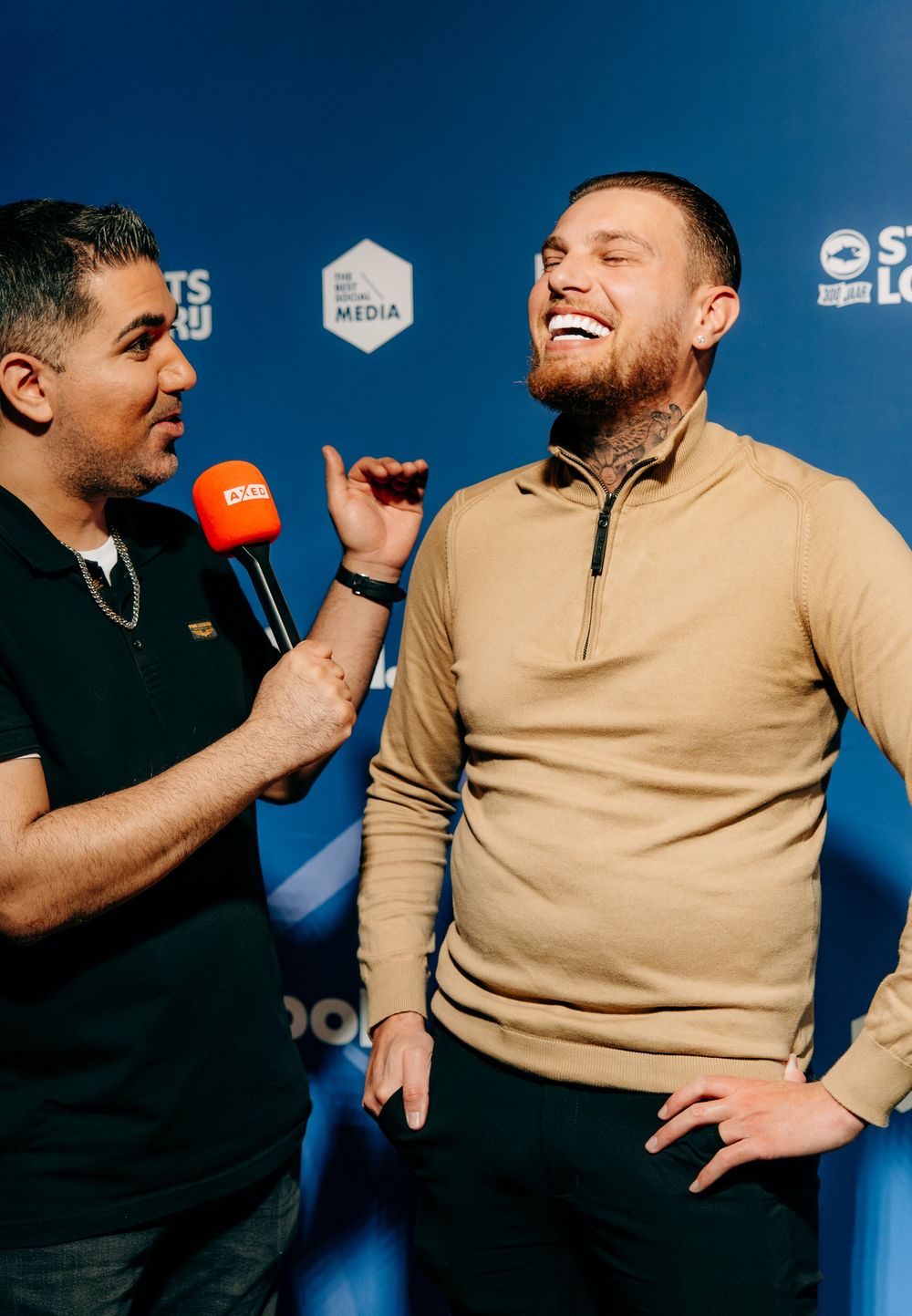 Two men laughing during lively interview with orange microphone at bol.com branded event backdrop.