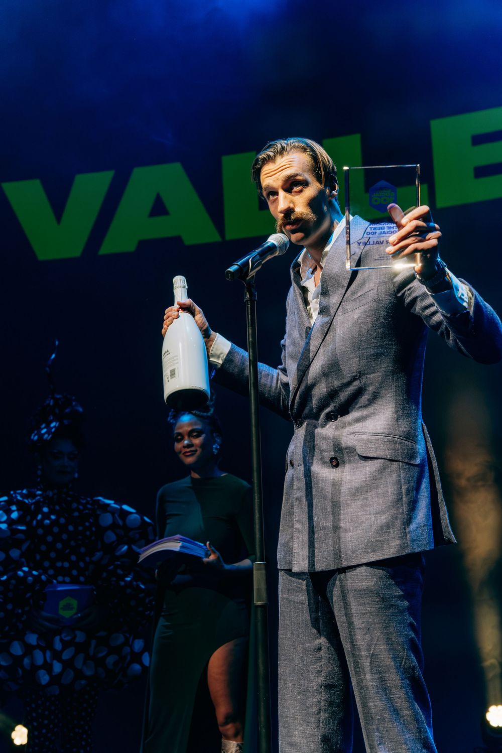 Man in grey suit holding champagne bottle and glass award on stage at awards ceremony.