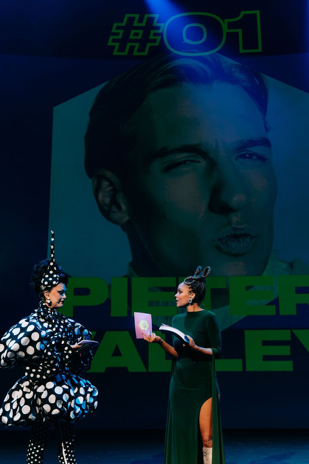 Two performers on stage holding books in front of a projected image reading Pieter Bailey #01 at a live event.