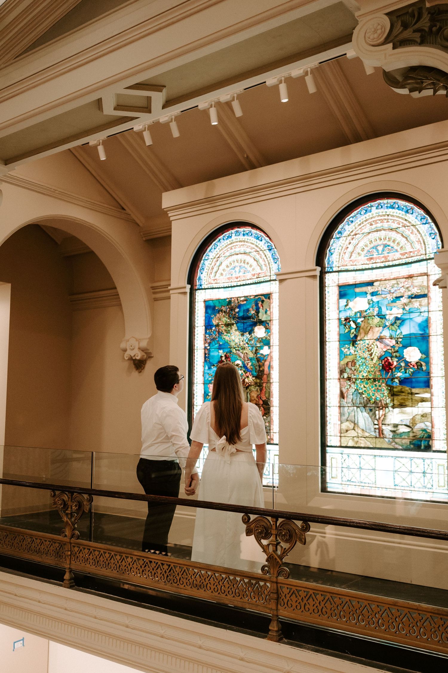 Couple holds hands while admiring stunning Tiffany stained glass windows in a museum gallery.