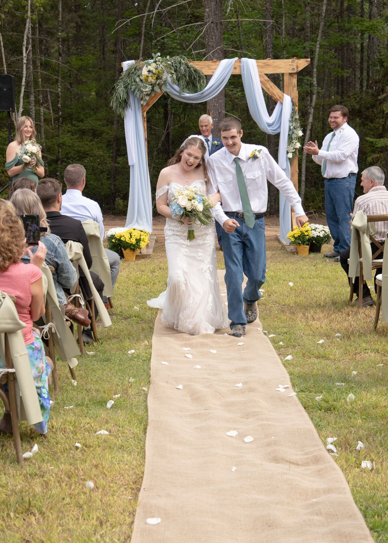 Outdoor wedding ceremony under wooden arch decorated with white fabric and greenery.
