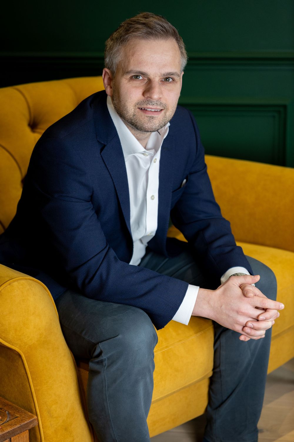 Professional portrait of someone in a navy suit sitting on a yellow couch against a dark green wall.