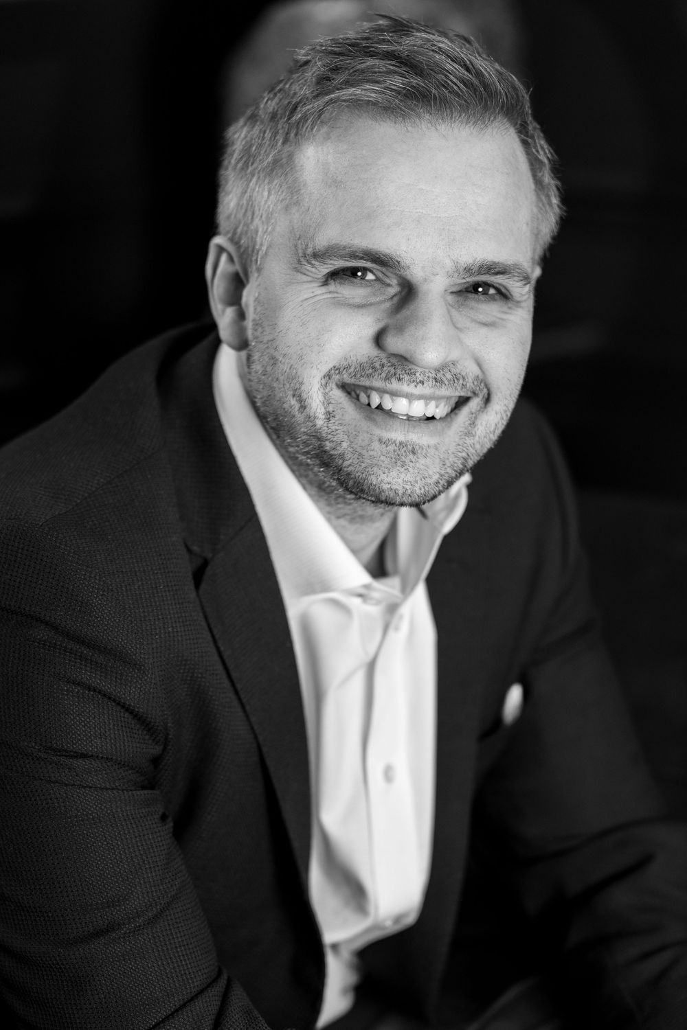 Professional black and white headshot portrait of a smiling man in a dark suit jacket and white dress shirt.