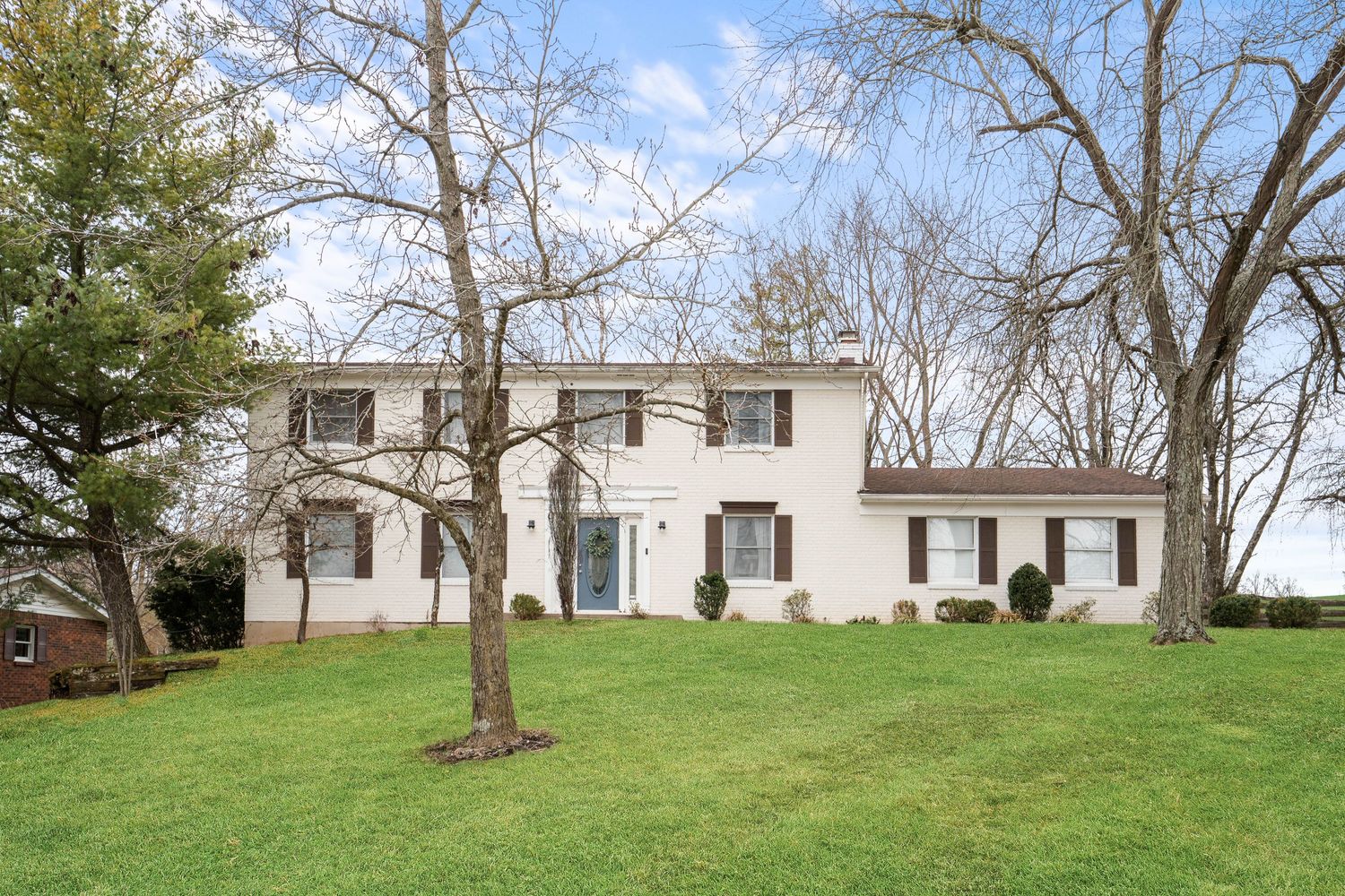 White two-story colonial home with brown shutters, green lawn, and bare trees on a cloudy spring day.