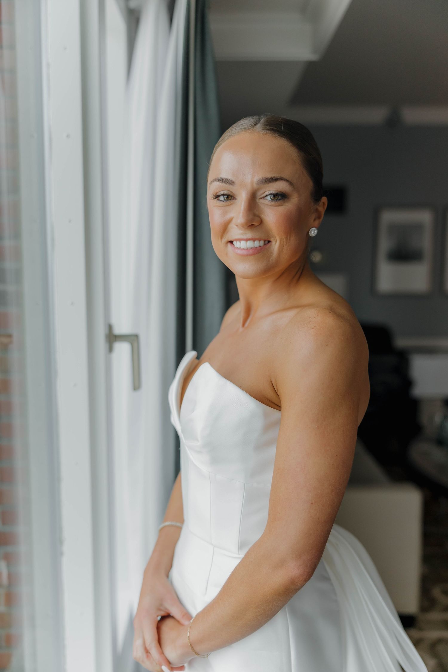 A bride in a strapless white wedding dress smiles while getting ready in a bright, window-lit room.