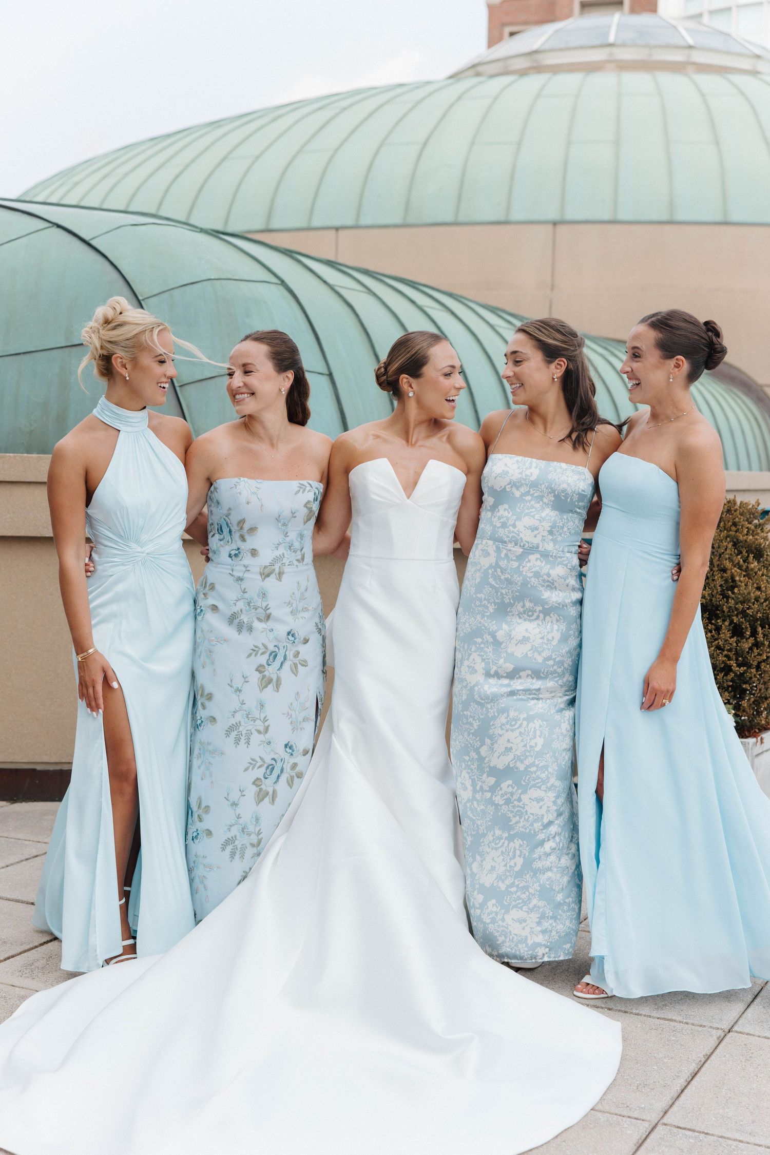 A bridal party wearing light blue bridesmaid dresses and a white wedding gown pose together outdoors.