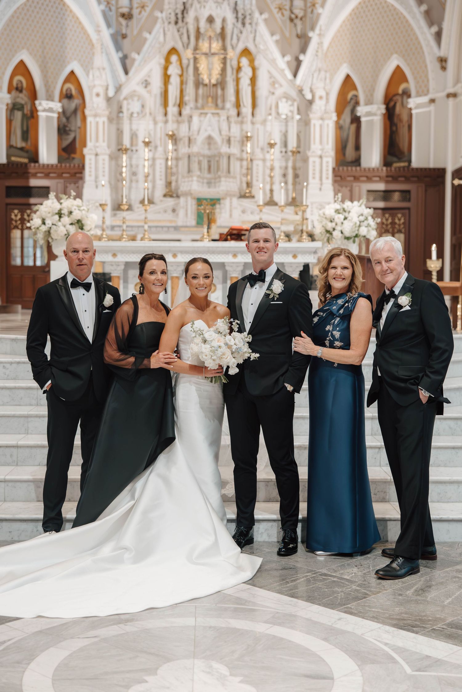 Elegant wedding party poses in formal attire inside ornate white church with gothic architecture and chandeliers.