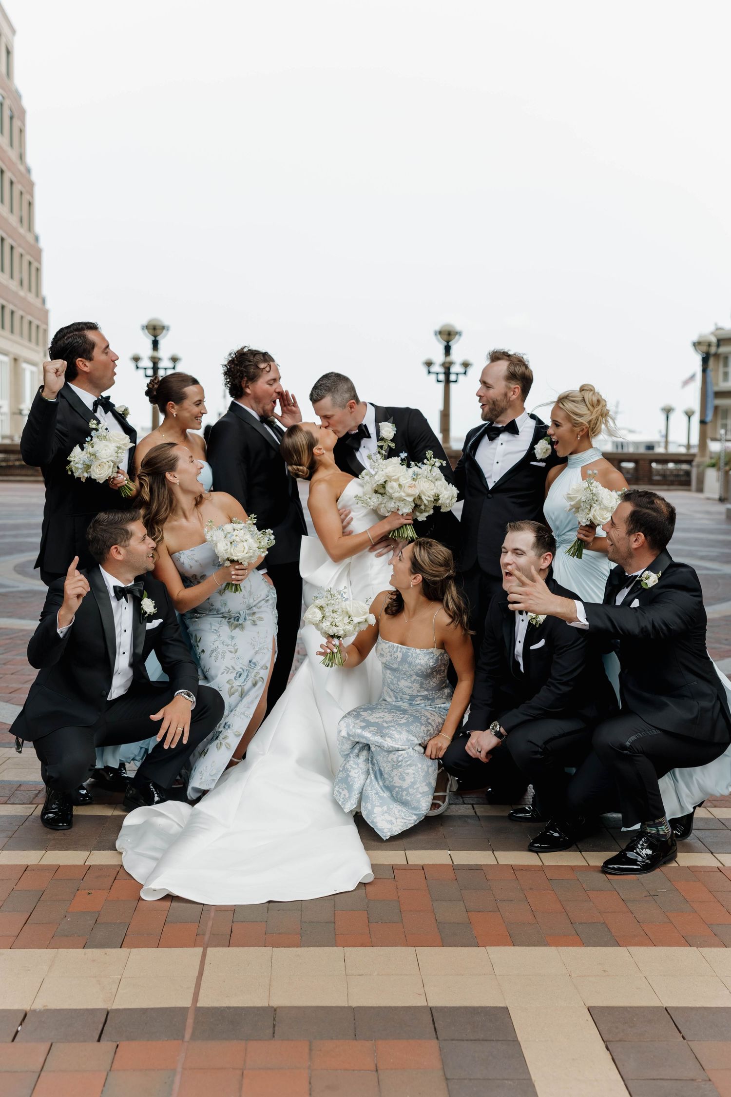 Wedding party in formal attire poses playfully on brick pavement while celebrating together.