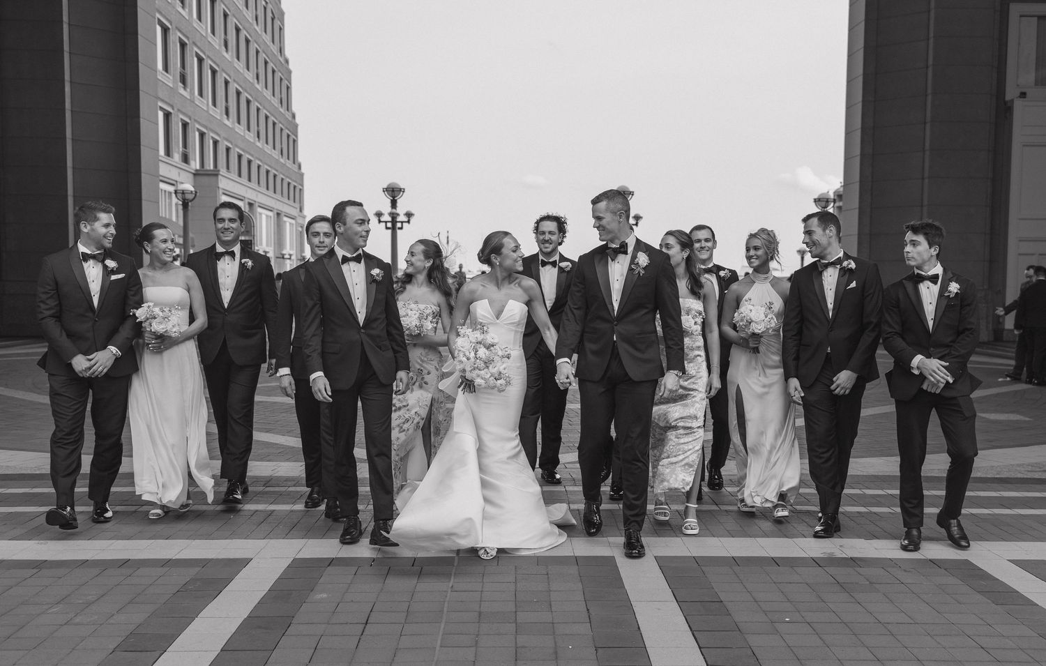 A wedding party walks together in formal attire down a paved walkway in black and white photography.