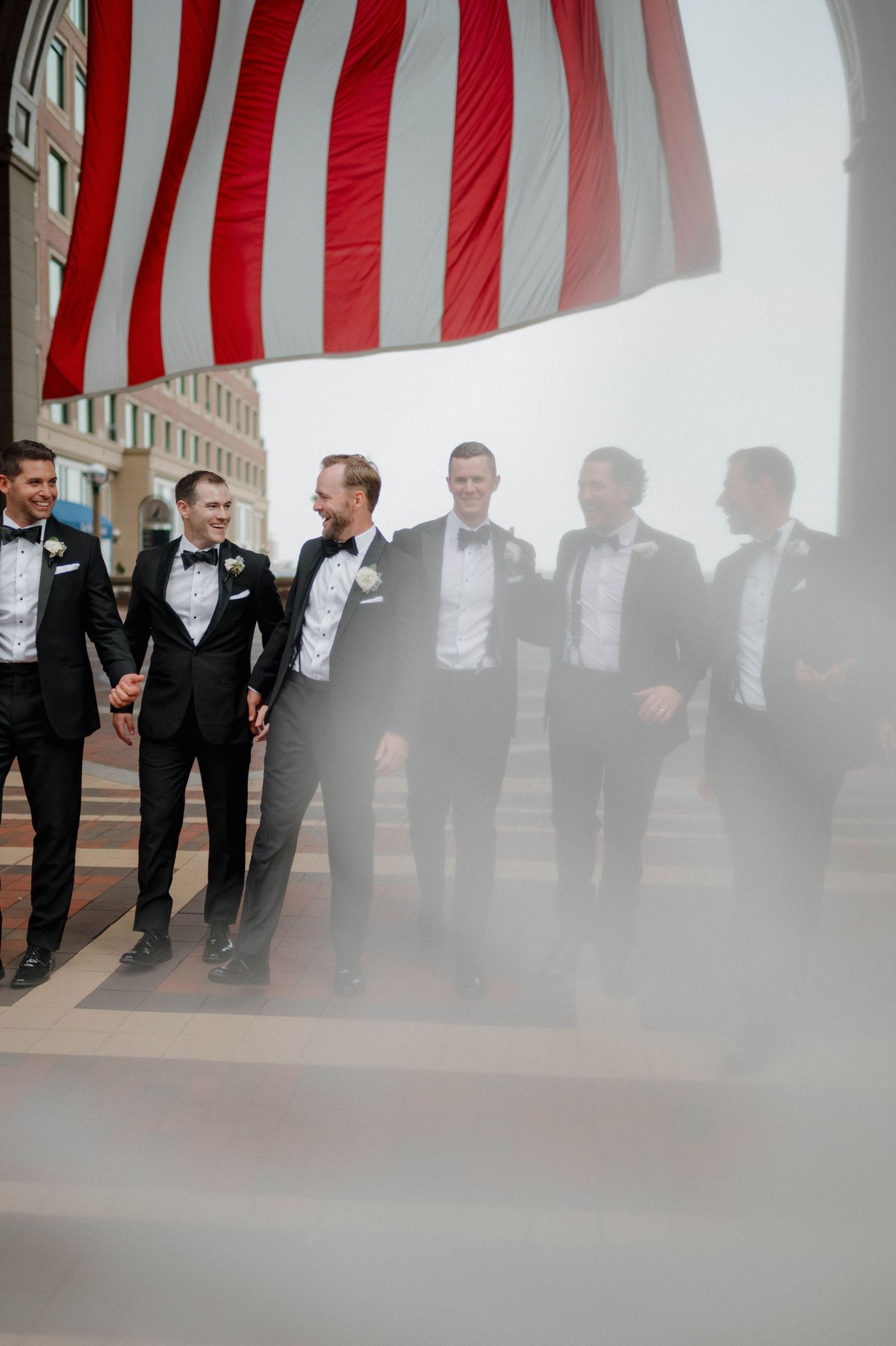 Groomsmen in black tuxedos stand in a row under American flags at an outdoor wedding venue.