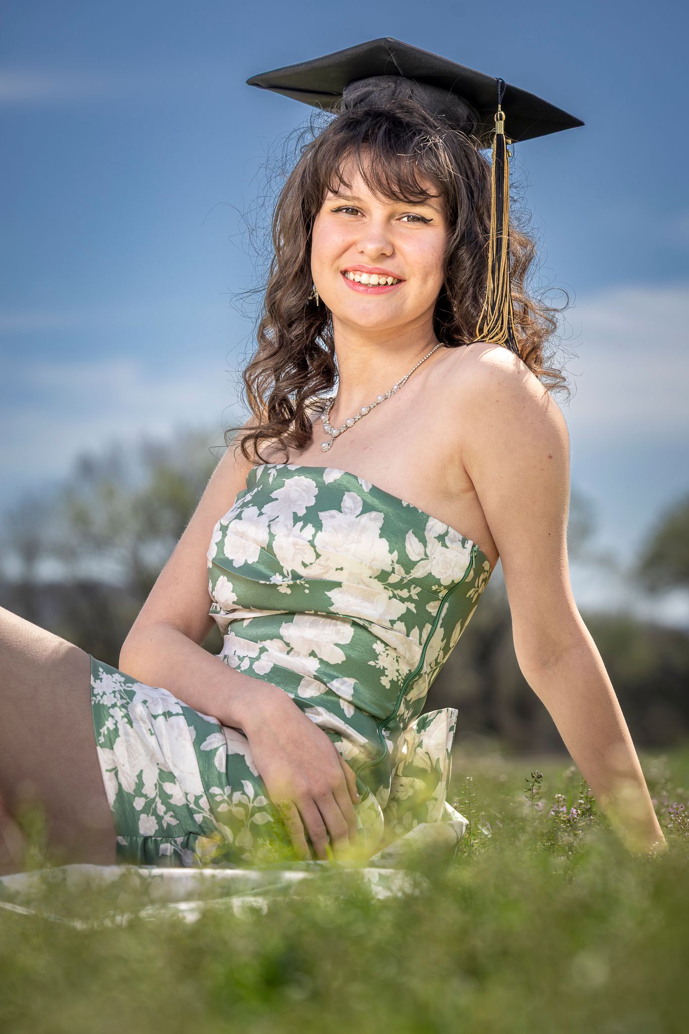 Graduate in floral dress poses in green grass field with graduation cap under blue sky.