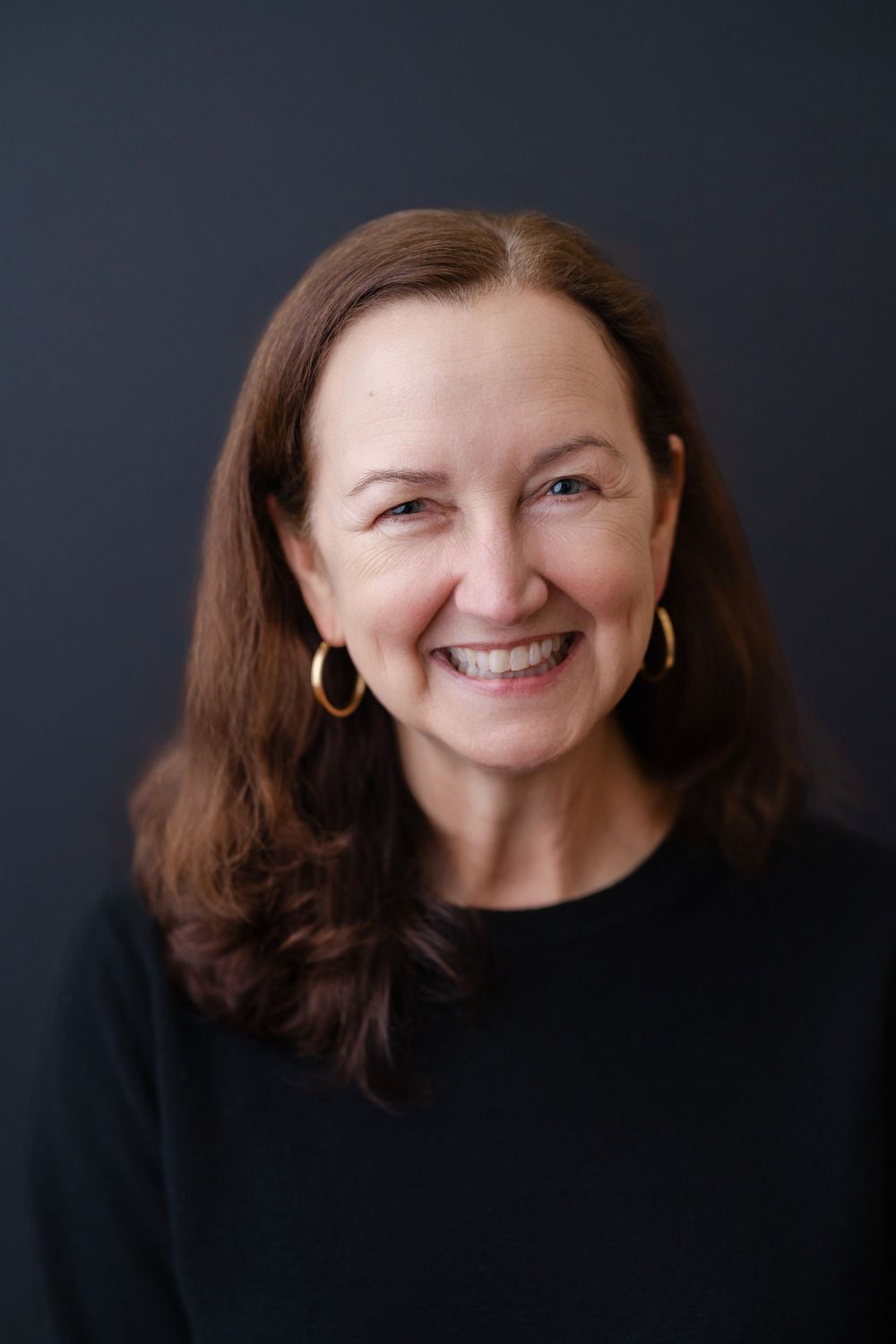Professional headshot of a woman with brown hair wearing a black sweater and gold hoop earrings against dark background.
