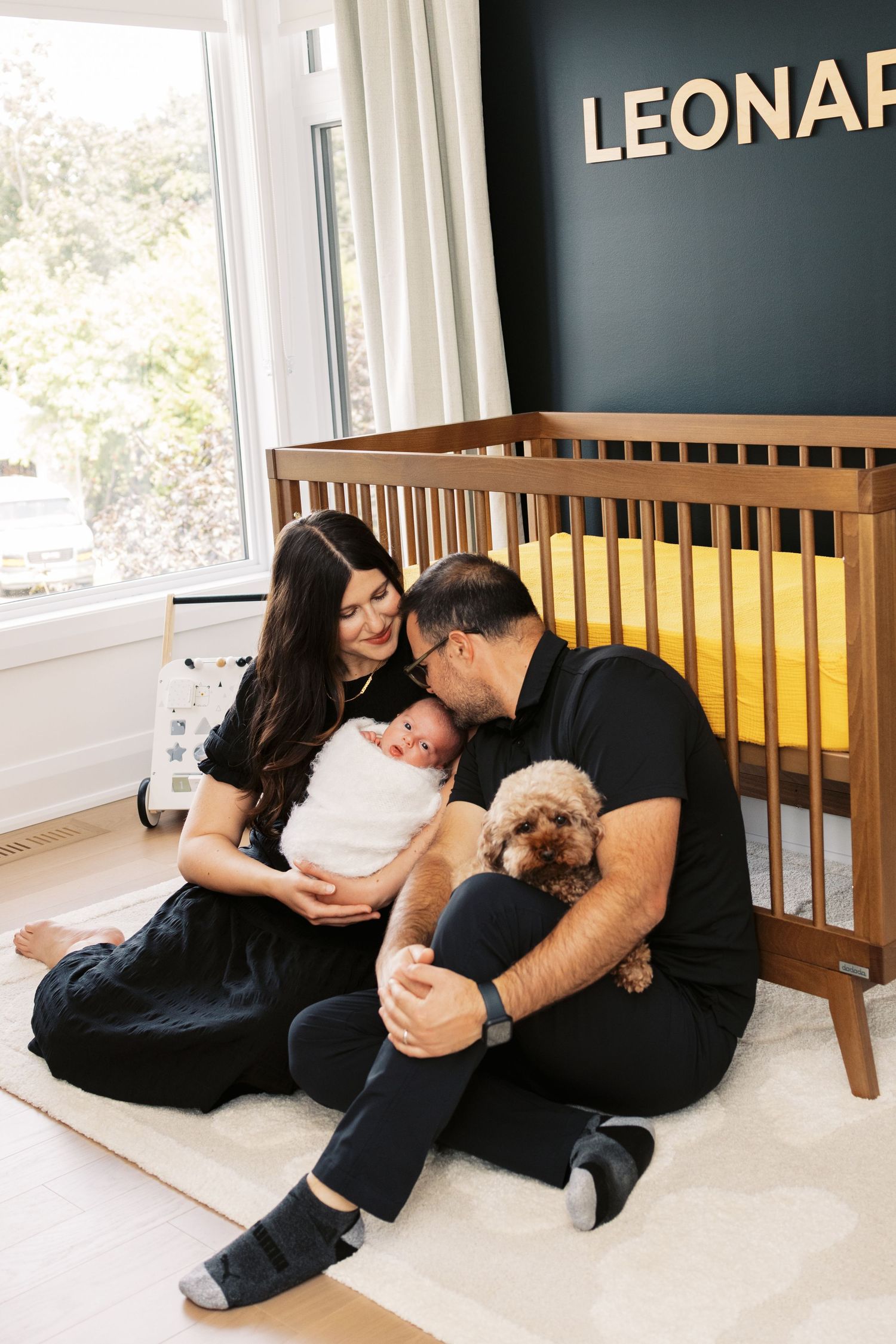New parents in black sit on nursery floor kissing newborn while holding small dog near wooden crib.