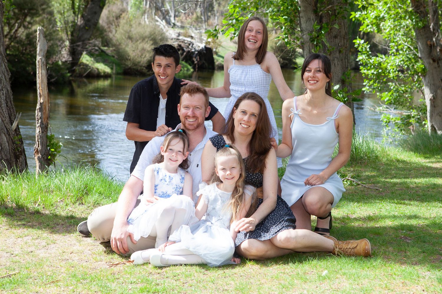 A family poses together outdoors in a natural setting with trees and dappled sunlight creating a warm atmosphere.