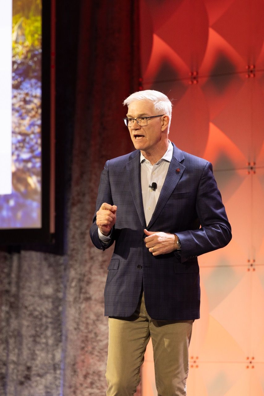A man and keynote speaker in a blue blazer and khaki pants speaks on stage at a conference with a red backdrop at an event in Dallas, TX