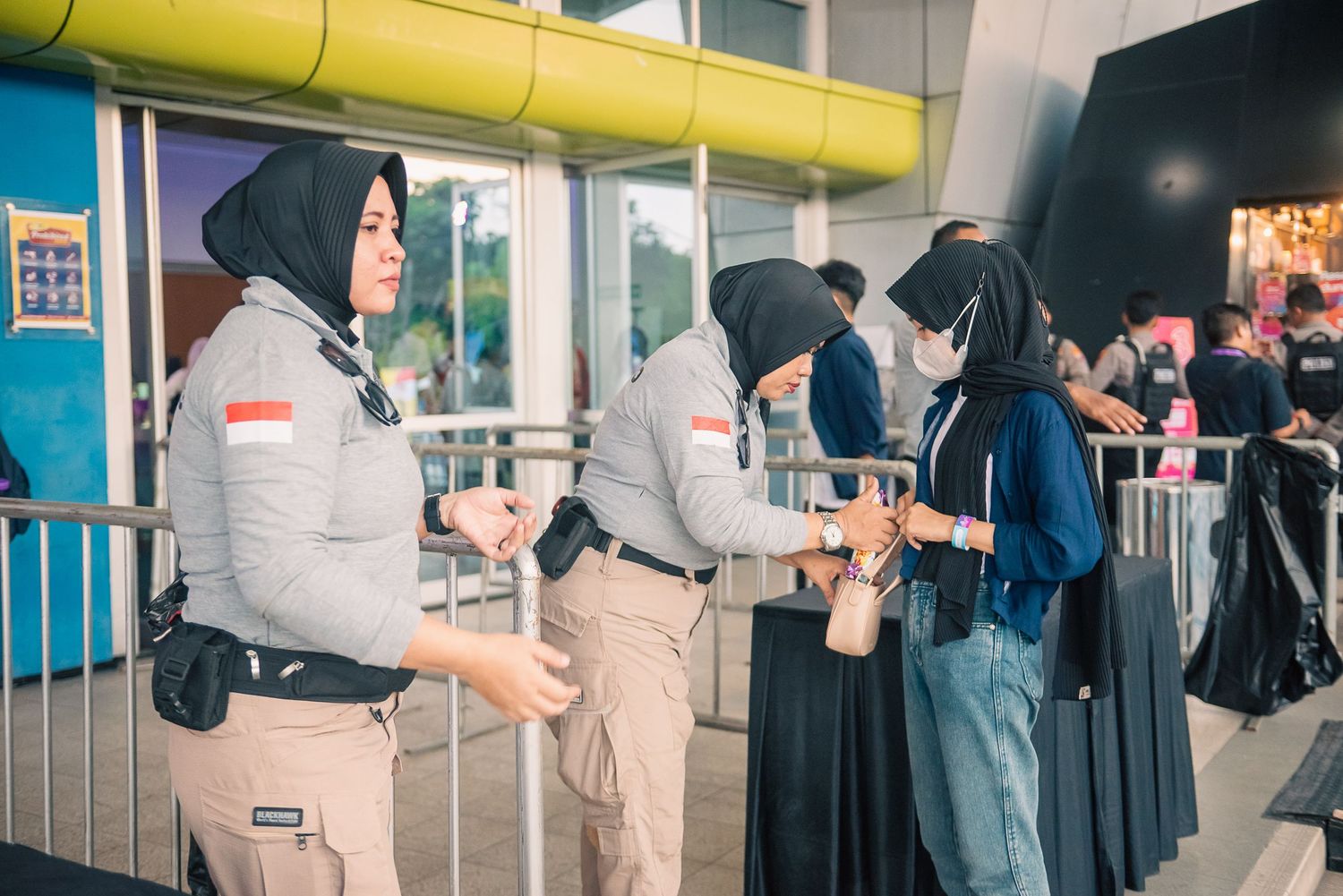 Female security guards in hijabs checking visitors at an outdoor event entrance with yellow and blue decor.